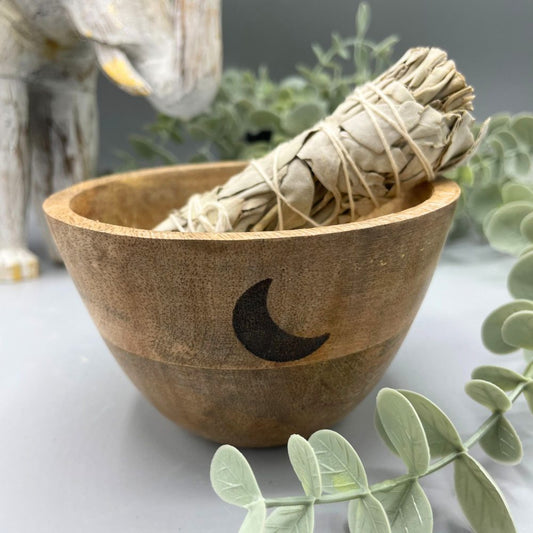 A bundle of tied sage rests in a small wooden bowl, which features a crescent moon. Greenery and a partial elephant statue surround the scene on a light gray surface.