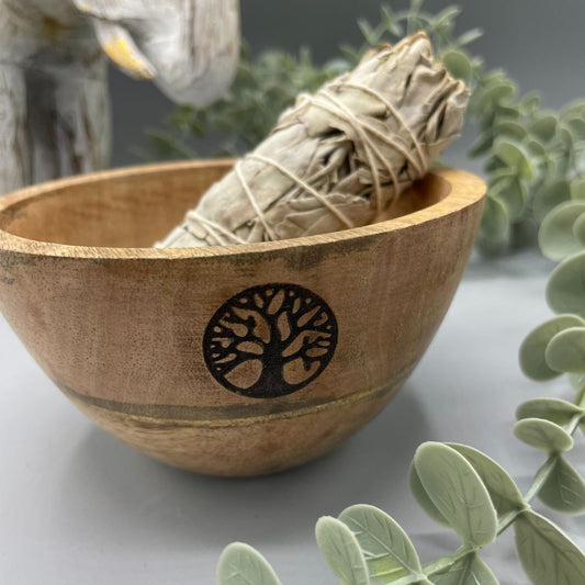 A tied sage bundle is positioned in a wooden bowl adorned with a tree design. The bowl sits on a gray surface, surrounded by greenery and a partial view of a white statue.