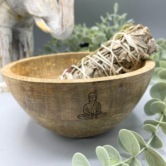 A bundle of sage rests inside a wooden bowl, etched with a seated Buddha. Greenery surrounds the bowl, which sits on a gray surface.