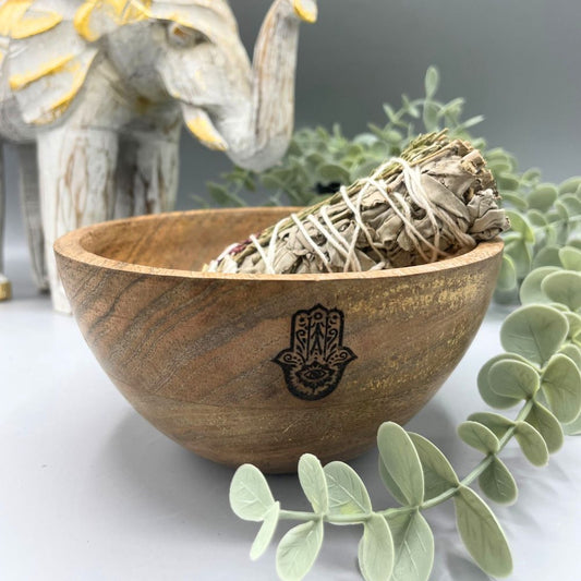 A wooden bowl with a Hamsa symbol etched on its side holds a sage bundle. The bowl is surrounded by green faux foliage against a gray background, with a decorative elephant in the background.