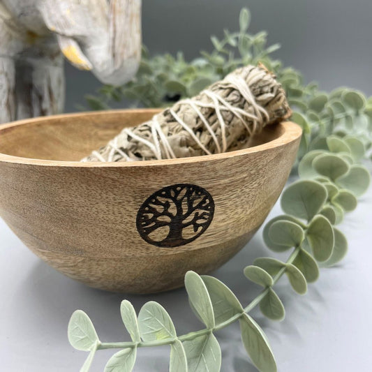 Sage bundle sits in wooden bowl with a tree design. Decorative greenery and elephant statue are in background on a gray surface.