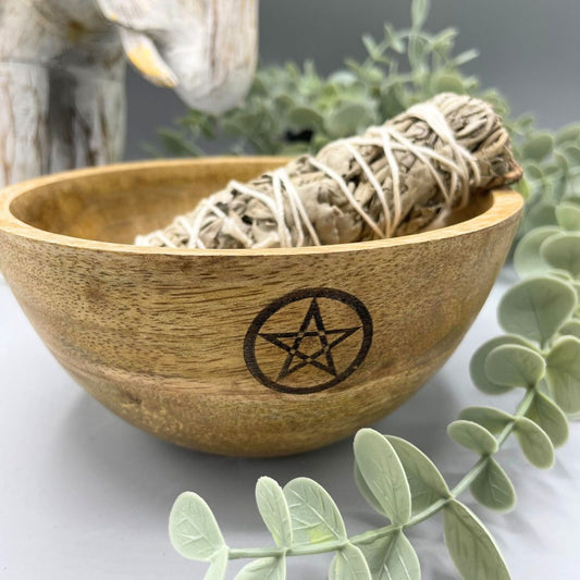 Sage rests in a wooden bowl with a pentagram symbol on its side, placed against a light grey backdrop with greenery and a statue visible in the background.