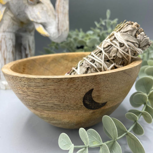 A sage bundle rests in a wooden bowl adorned with a crescent moon. Greenery and an elephant statue are blurred in the background.