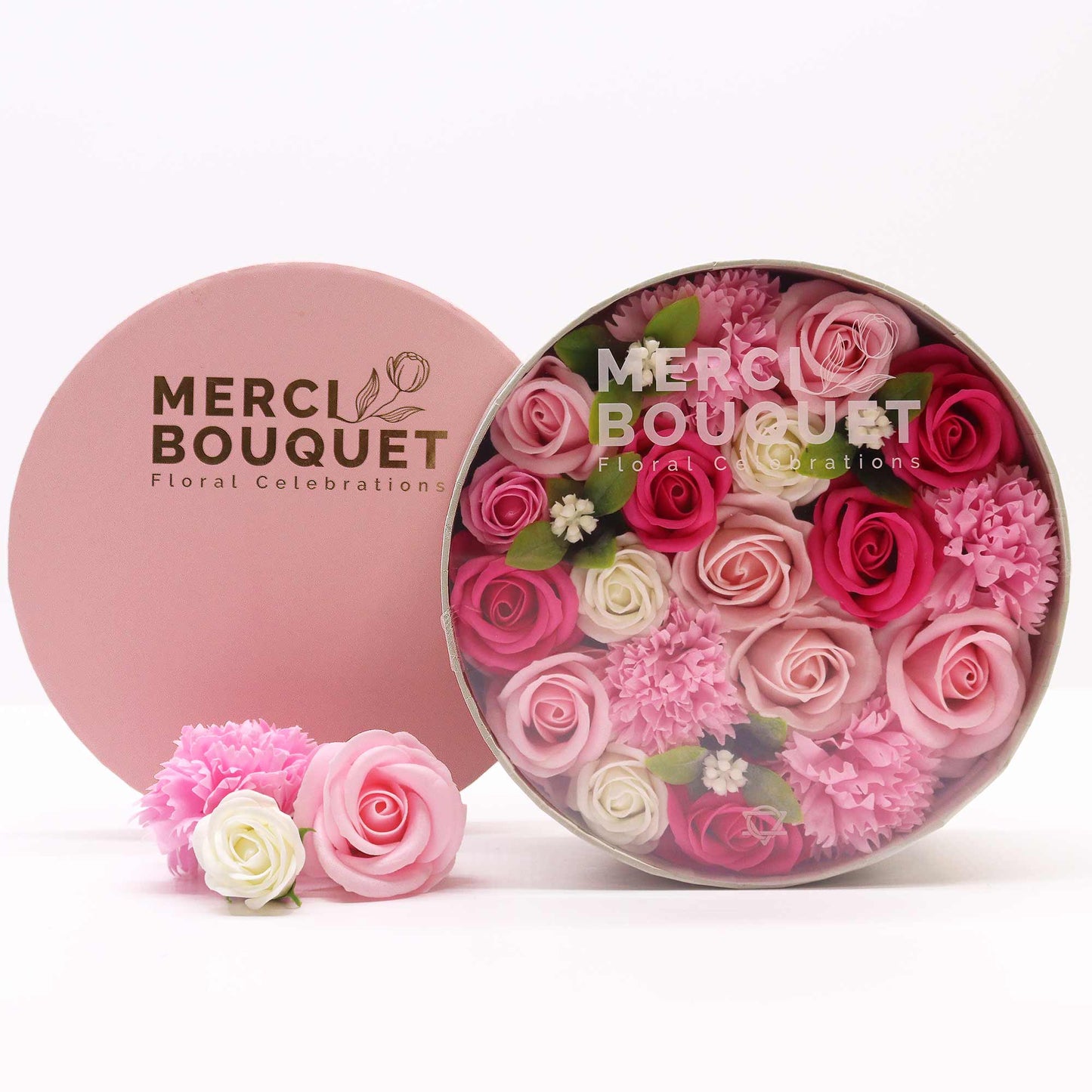 A round box containing an arrangement of pink and white soap roses and carnations sits on a white surface. Three additional soap flowers are beside the box. The box and lid are pink and display the logo: "MERCI BOUQUET Floral Celebrations."