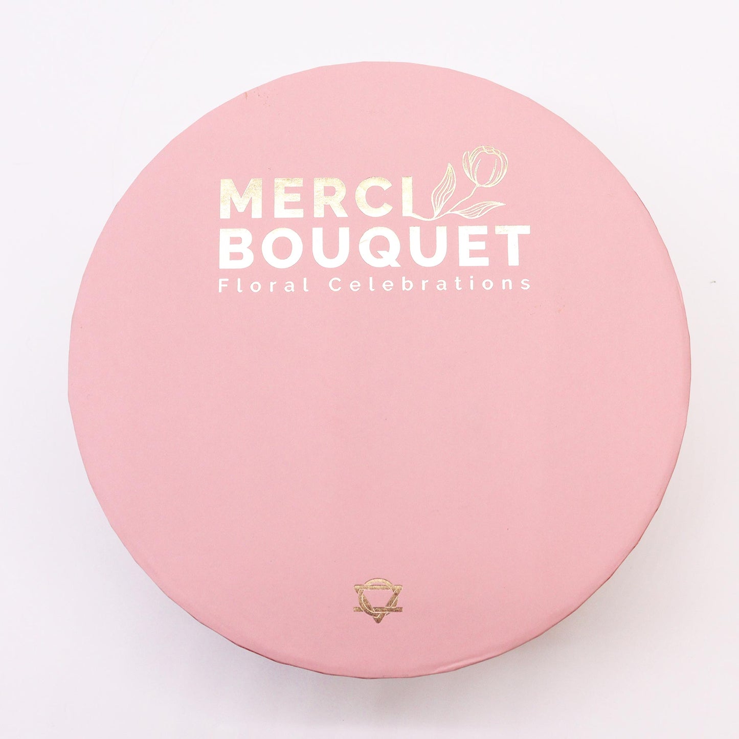 A pink round box sits on a white background. It displays "MERCI BOUQUET Floral Celebrations" in gold and white lettering. A small gold logo is at the bottom.