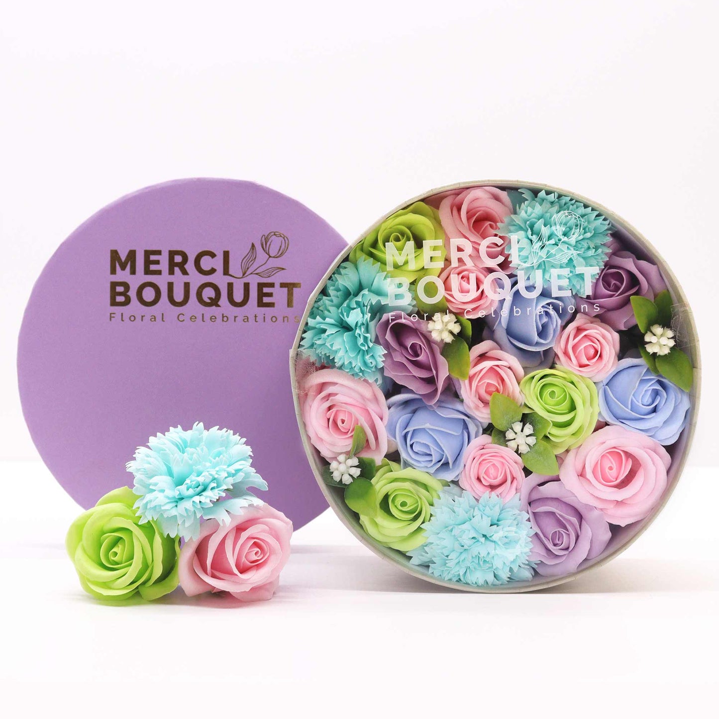 A pastel soap flower arrangement sits in a round box. A smaller arrangement is beside it. Both are on a white surface. The box and a separate lid are branded "MERCI BOUQUET Floral Celebrations".