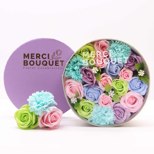 A pastel soap flower arrangement sits in a round box. A smaller arrangement is beside it. Both are on a white surface. The box and a separate lid are branded "MERCI BOUQUET Floral Celebrations".