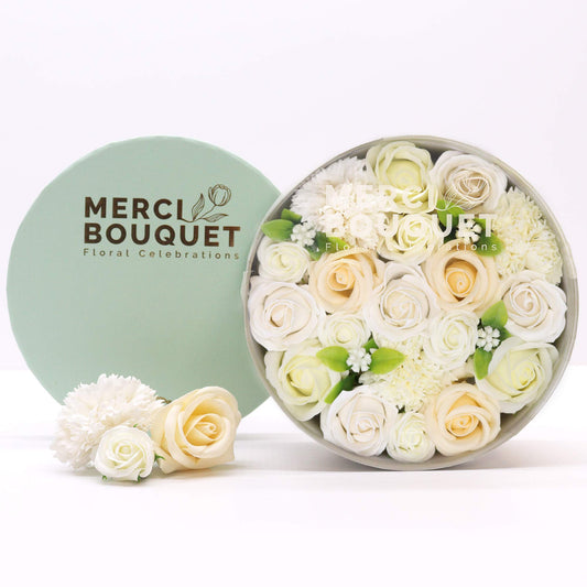 A circular arrangement of cream and white soap roses and other flowers sits inside a gray box. It rests next to a light green box lid with “MERCI BOUQUET Floral Celebrations” printed on it. Three additional soap flowers are placed near the box.