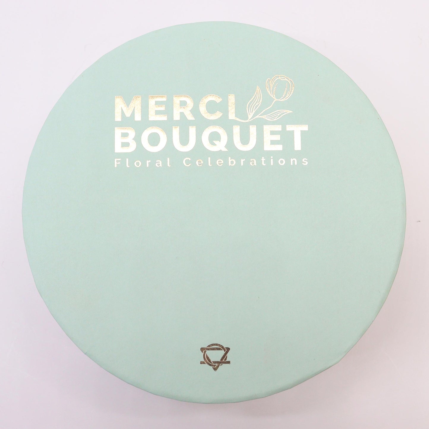 A round, light-green box sits on a white surface. The box displays "MERCI BOUQUET Floral Celebrations" in gold lettering, along with a small floral design and a logo.