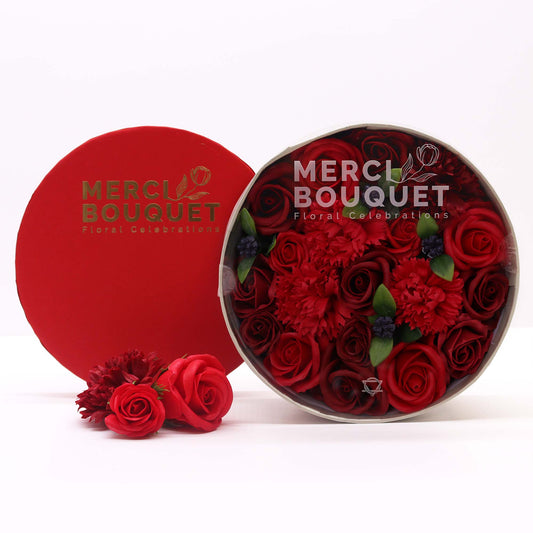 A red rose bouquet, arranged in a round box, sits on a white surface. The box is labeled “MERCI BOUQUET Floral Celebrations.” A smaller bouquet of similar roses rests beside the larger arrangement.