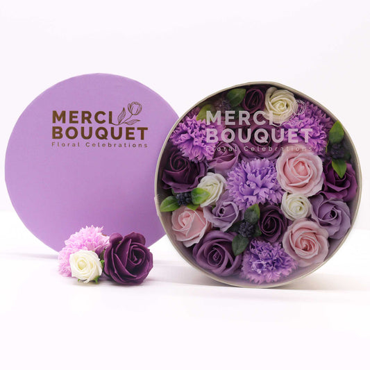 A round box containing a bouquet of purple, pink, and white soap roses and carnations sits on a white surface next to a smaller, matching box with a few additional flowers. The boxes are labeled "MERCI BOUQUET Floral Celebrations."