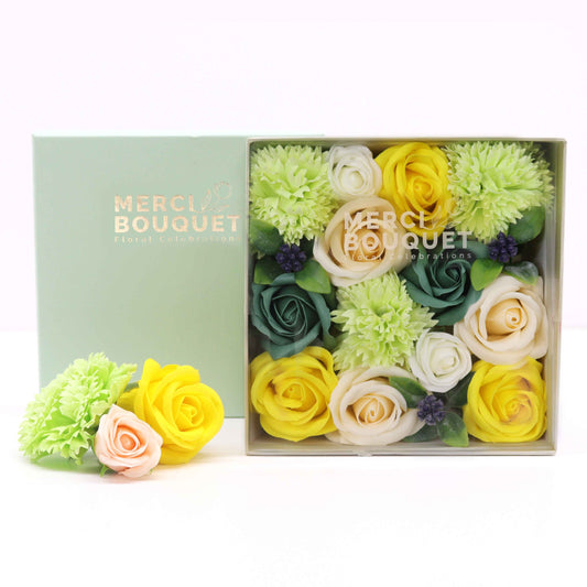 A soap flower bouquet, arranged in a light green box, sits on a white surface. The box's label reads: "MERCI BOUQUET Floral Celebrations". A smaller, similar arrangement is positioned to the left.