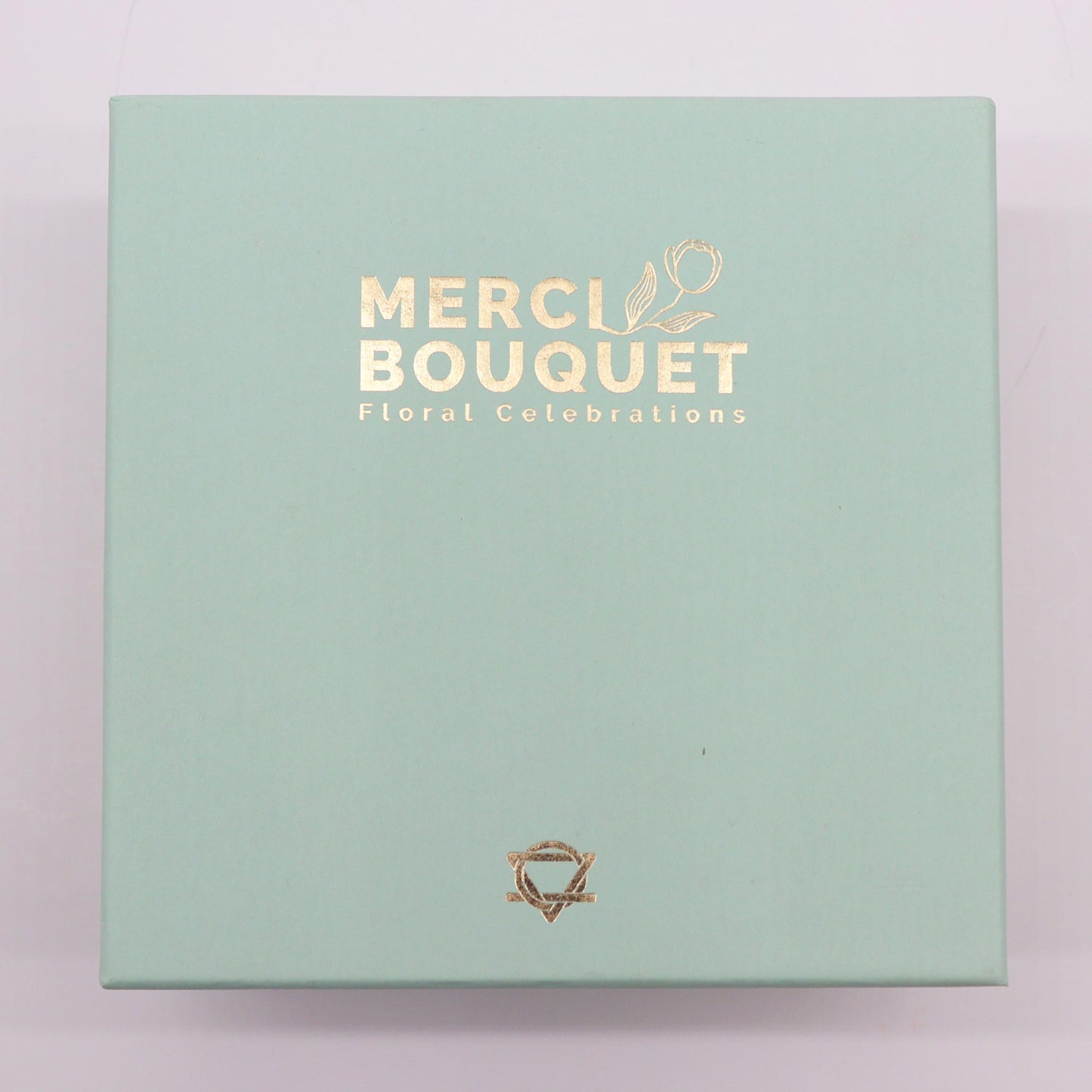 A light-green box rests on a surface. It displays gold lettering: "MERCI BOUQUET Floral Celebrations" and a small gold logo.