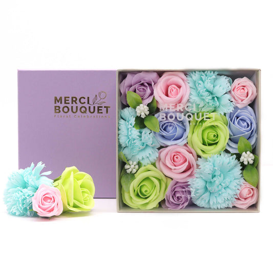 A box of pastel soap flowers sits on a white surface; roses and carnations are arranged inside a lavender box. The box is labeled "MERCI BOUQUET Floral Celebrations". A smaller grouping of soap flowers sits outside the box.