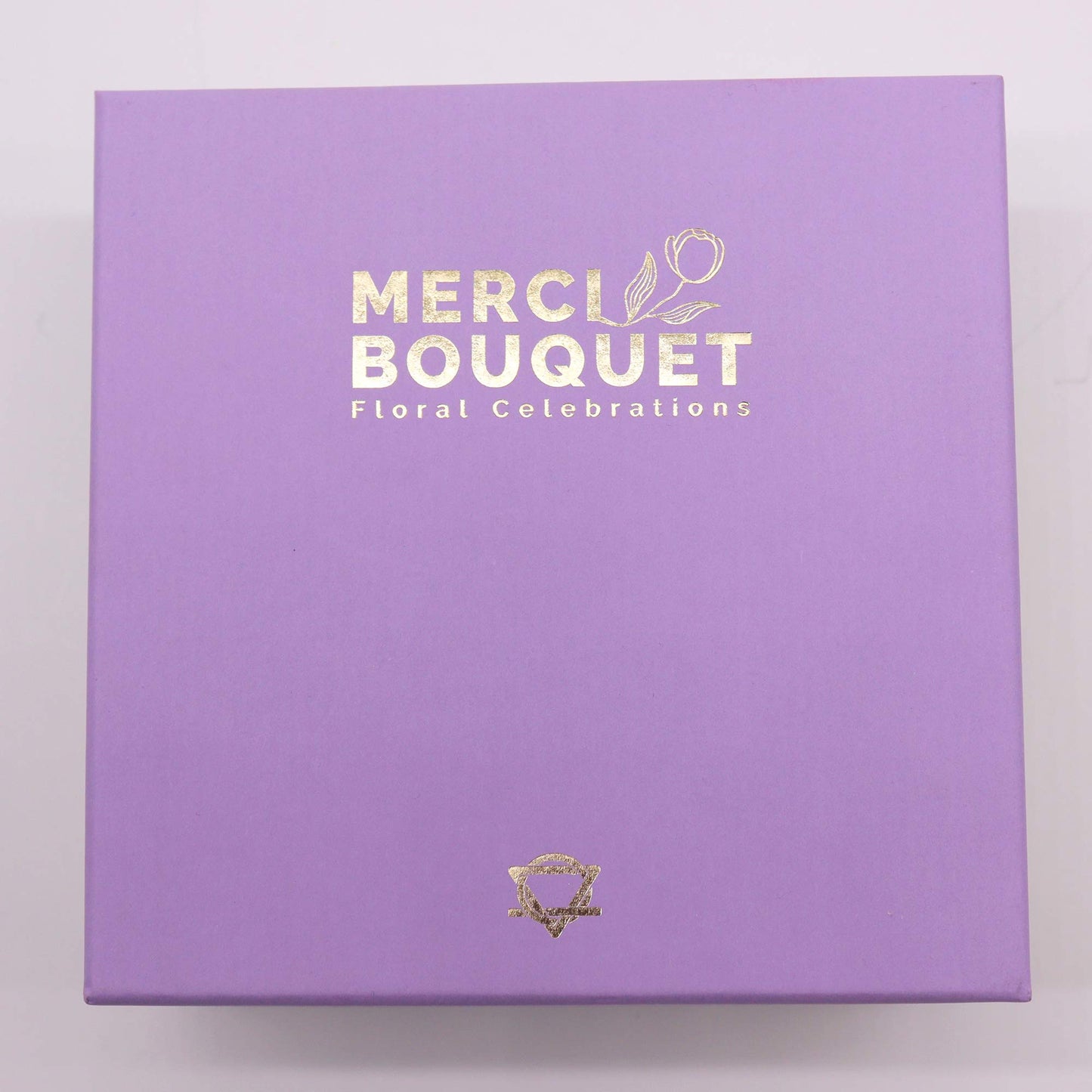 A lavender box sits on a white surface. The box displays gold lettering: "MERCI BOUQUET," and "Floral Celebrations." A small gold logo is at the bottom.
