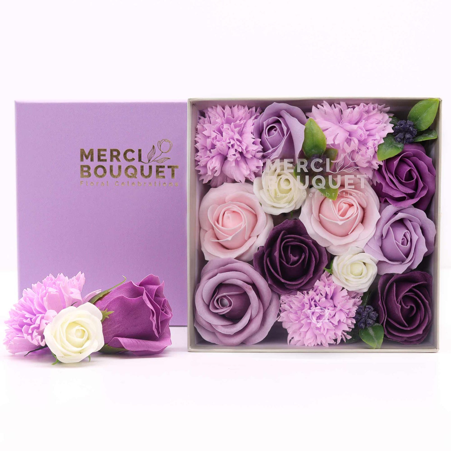 A square box contains a bouquet of artificial roses and carnations in shades of purple, pink, and white. The box is lavender, displaying "MERCI BOUQUET Floral Celebrations". A smaller arrangement rests beside it.