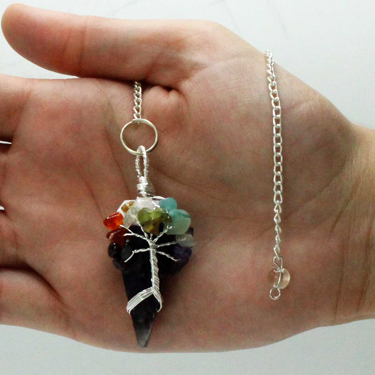 A silver-wired, cone-shaped crystal pendant with colorful stones is held on an open palm. A silver chain hangs from the pendant. The background is a blurry white.