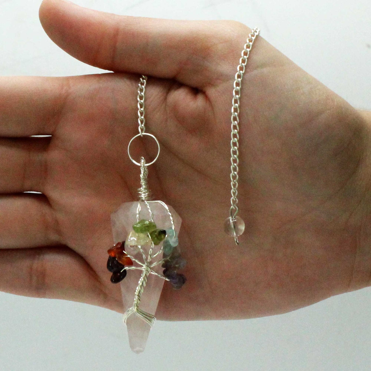 A hand holds a rose quartz crystal pendulum. The pendulum has a silver chain and tree of life made of colored gems, over a white background.