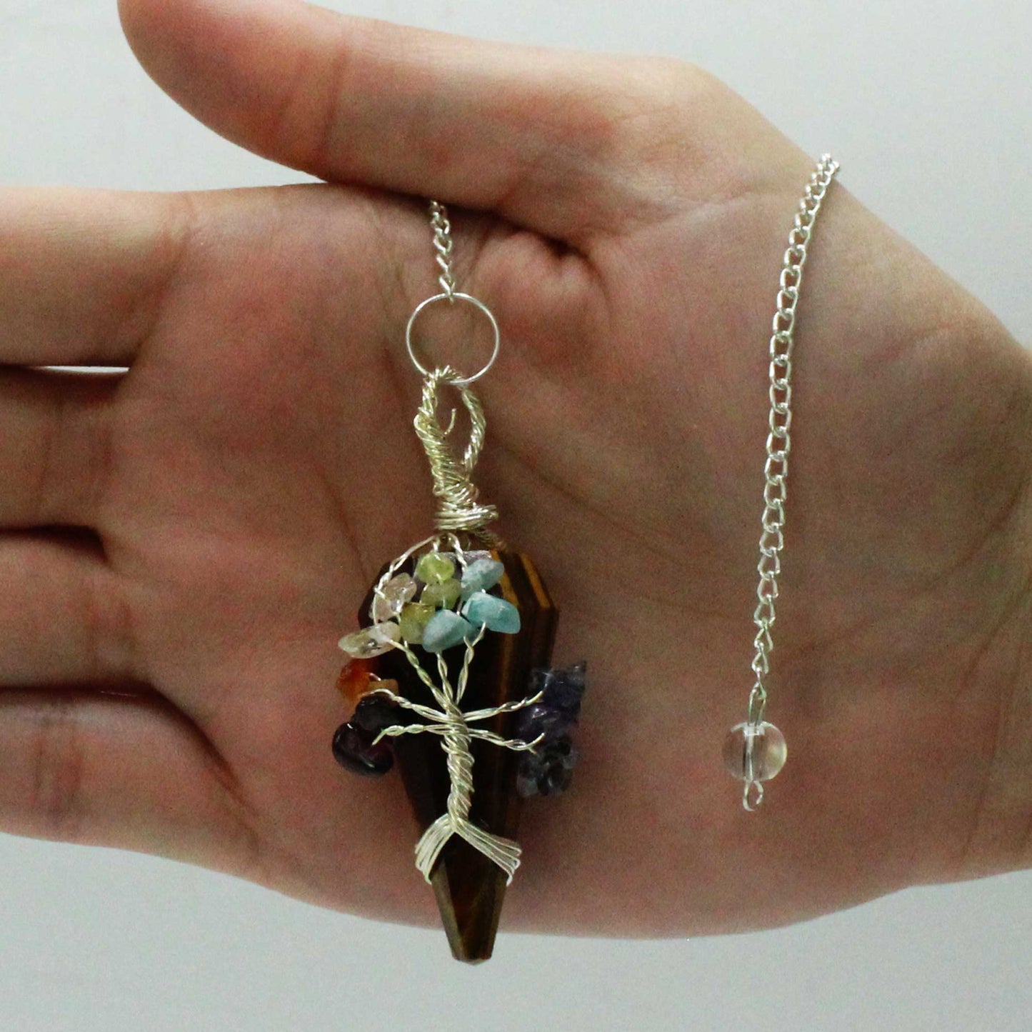 A tiger-eye pendant is held in a hand; silver wire in a tree shape holds colorful stones. A silver chain with a clear bead hangs to the right. The background is plain white.