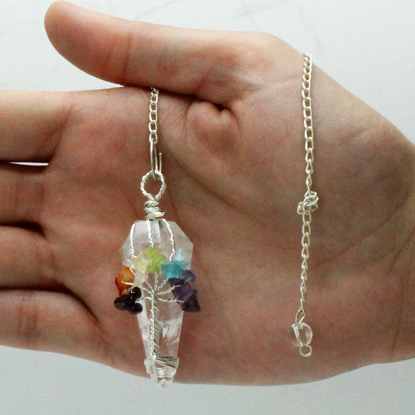 A hand holds a crystal pendant with a silver chain. The pendant is wrapped in silver wire with rainbow-colored stones arranged around it. The background is plain white.