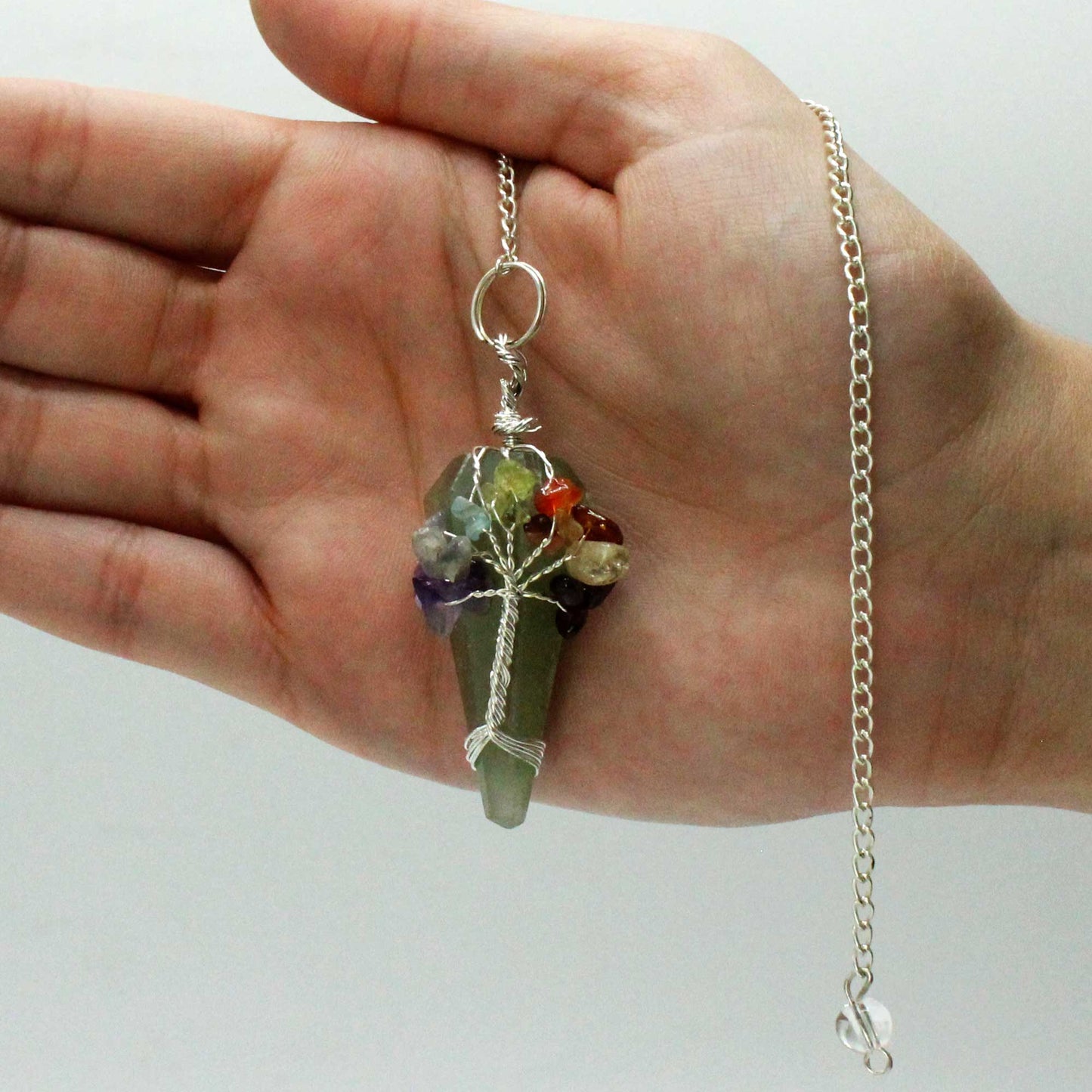 A hand holds a crystal pendulum. A silver chain is attached, and colorful stones are wired to the top, resembling a tree. The background is plain white.