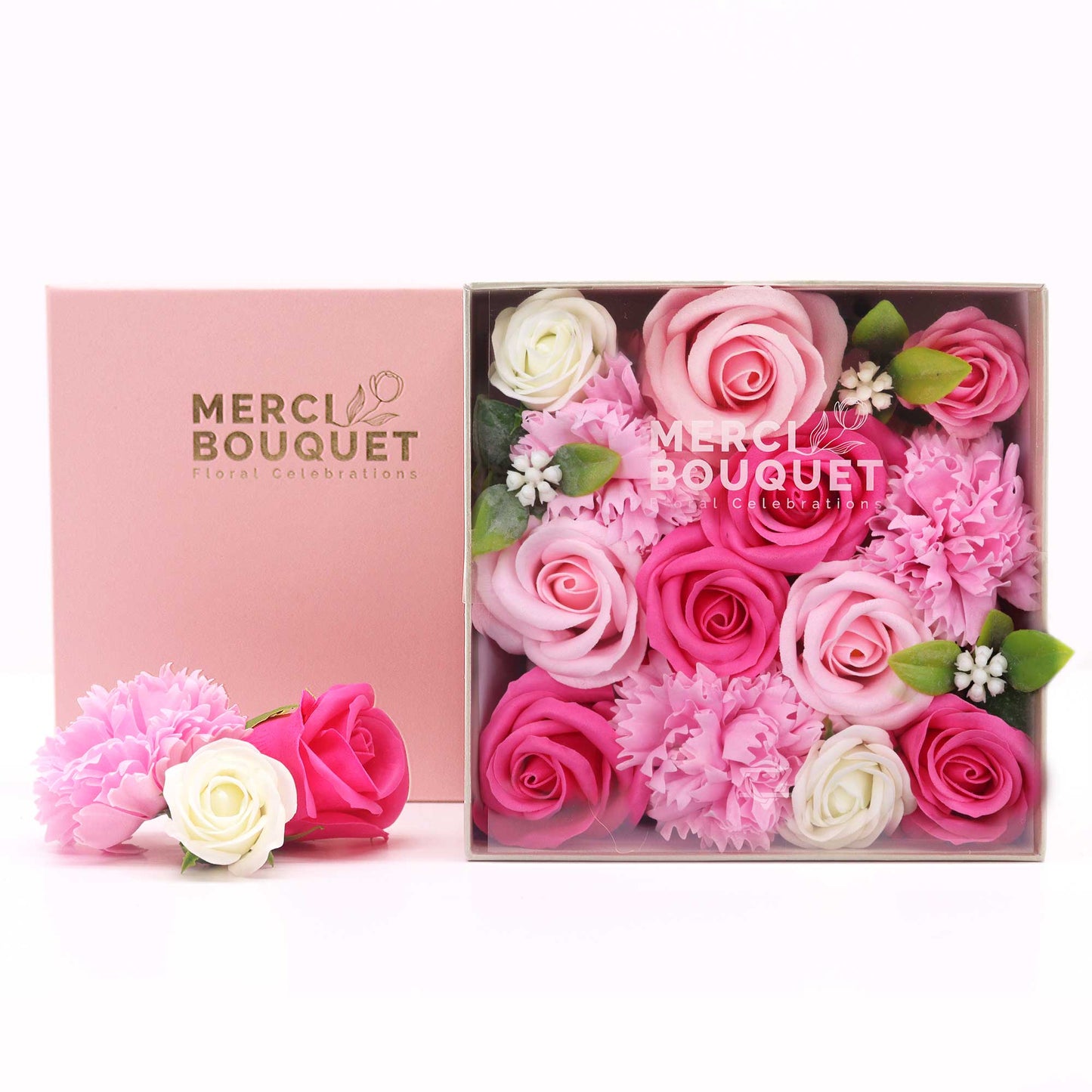 A pink box containing an arrangement of pink, white, and light pink soap roses and carnations sits on a white surface; a smaller arrangement of similar soap flowers rests beside it. The box is labeled "MERCI BOUQUET Floral Celebrations".