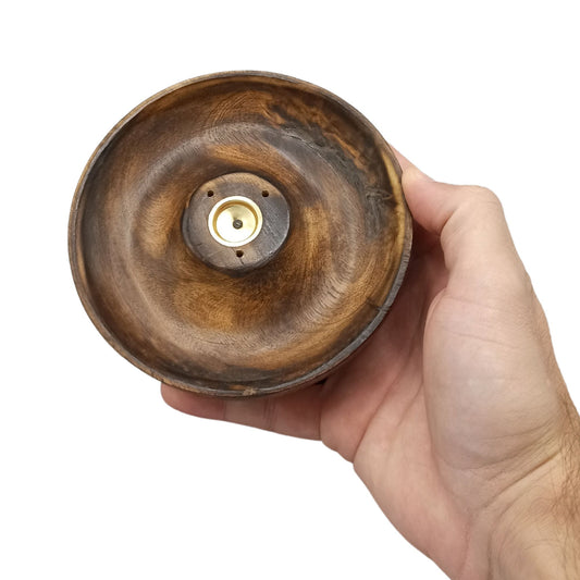 A hand holds a carved wooden bowl with a smooth, swirling pattern, featuring a central brass component and two small holes, against a plain white background.