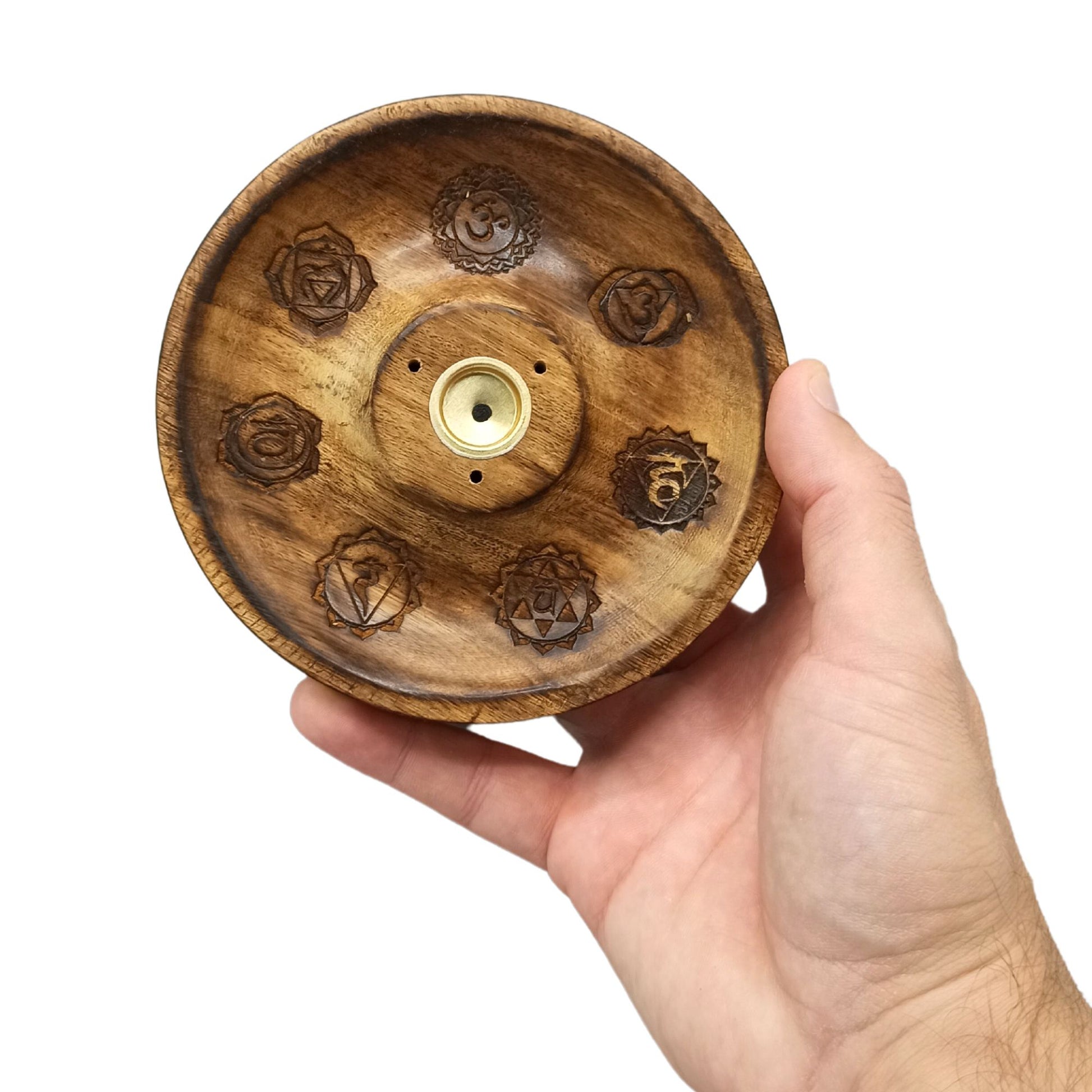 A hand holds a round wooden incense holder engraved with chakra symbols, featuring a central brass inlay with multiple holes, viewed against a plain white background.