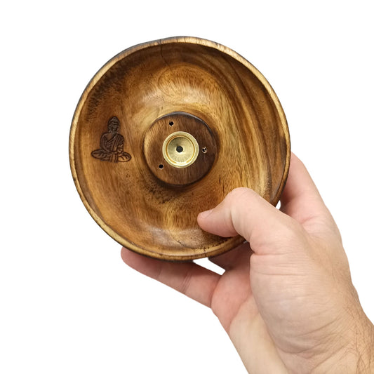 A wooden incense holder is being held in a hand. It features a carved Buddha figure and a central brass insert with several holes, against a plain white background.