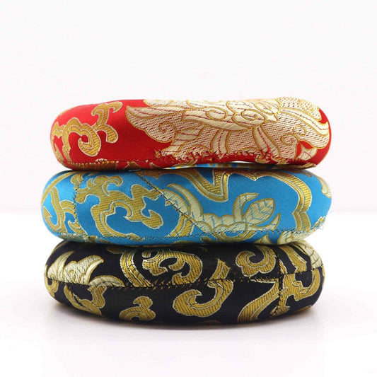 Three ornate, ring-shaped cushions, stacked vertically, display gold designs on red, turquoise, and black fabric. The cushions rest on a white surface.