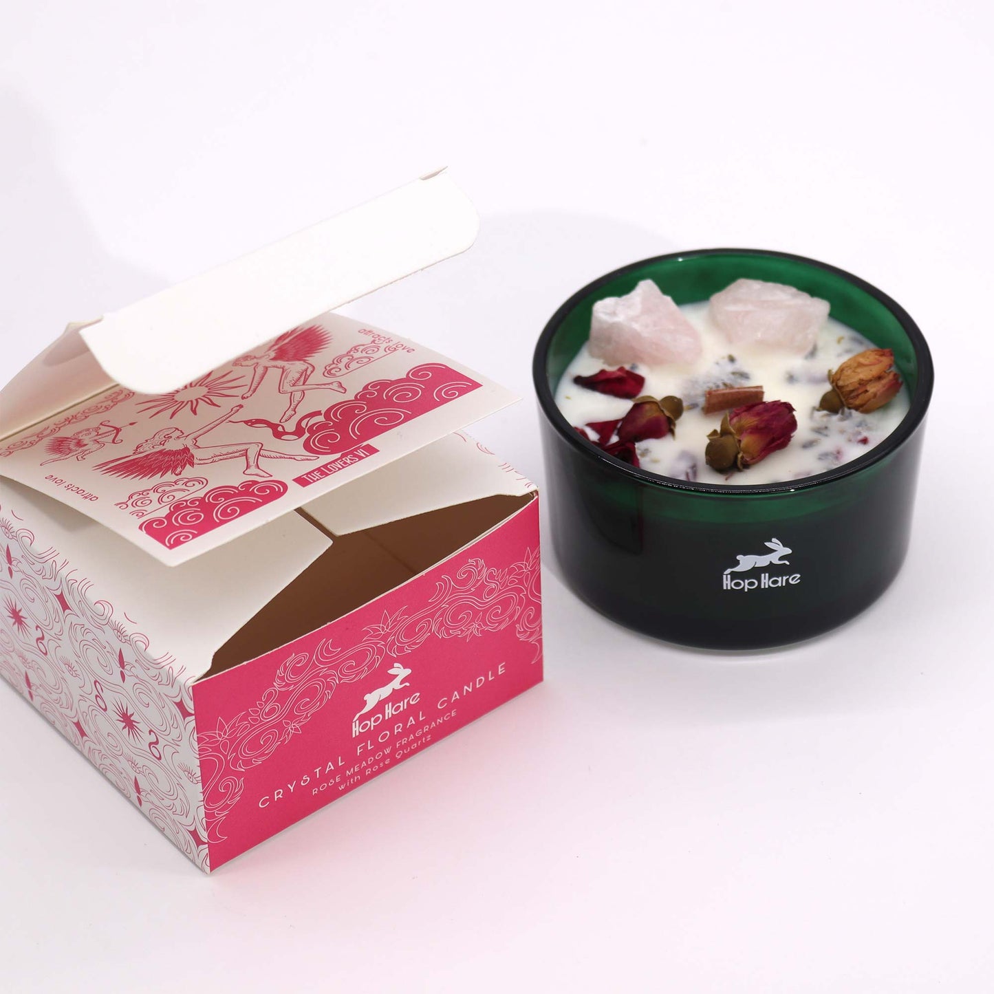 A partially opened "Hop Hare Crystal Floral Candle" box sits next to the candle, which contains chunks of rose quartz and dried rose petals in white wax in a green holder. Box reads, "THE LOVERS VI", and, "attracts love."