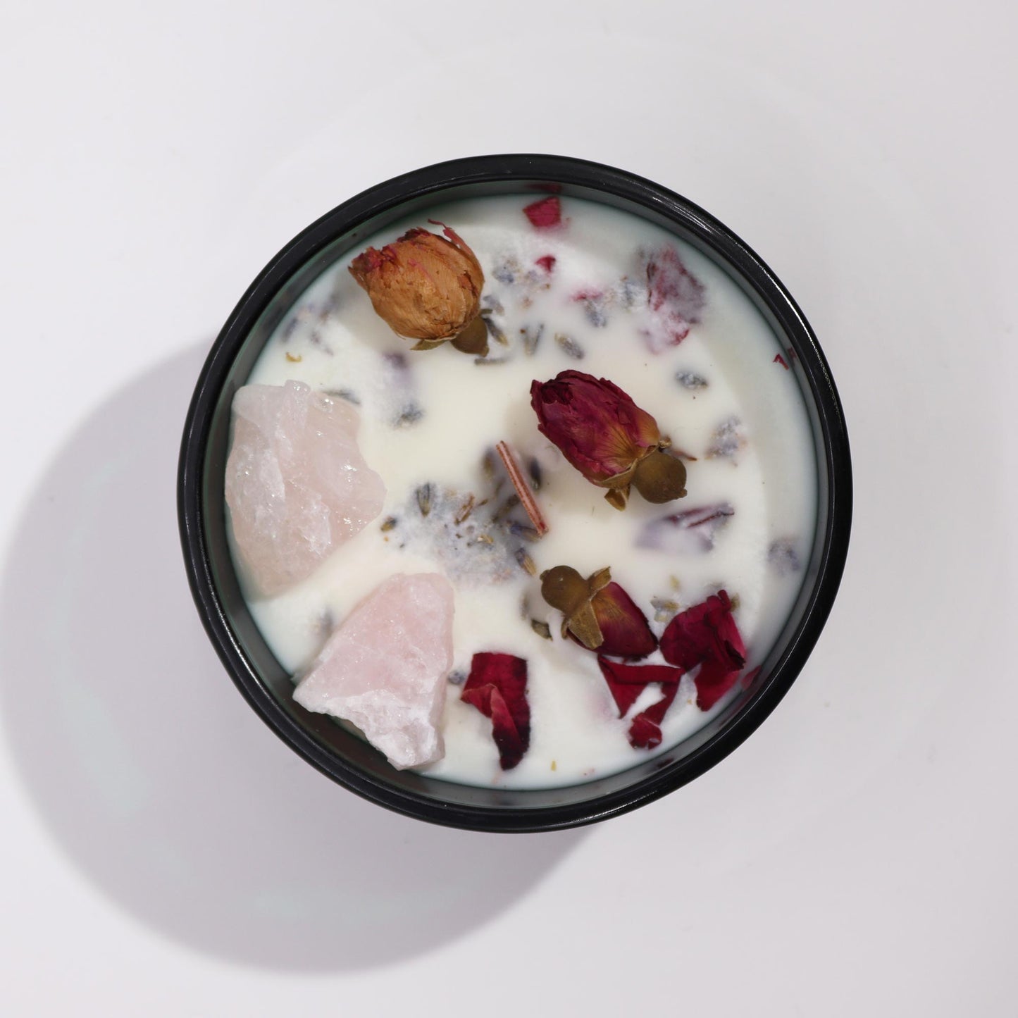 A round black candle holder contains wax, rose buds, and rose quartz chunks. The composition sits on a plain white surface.