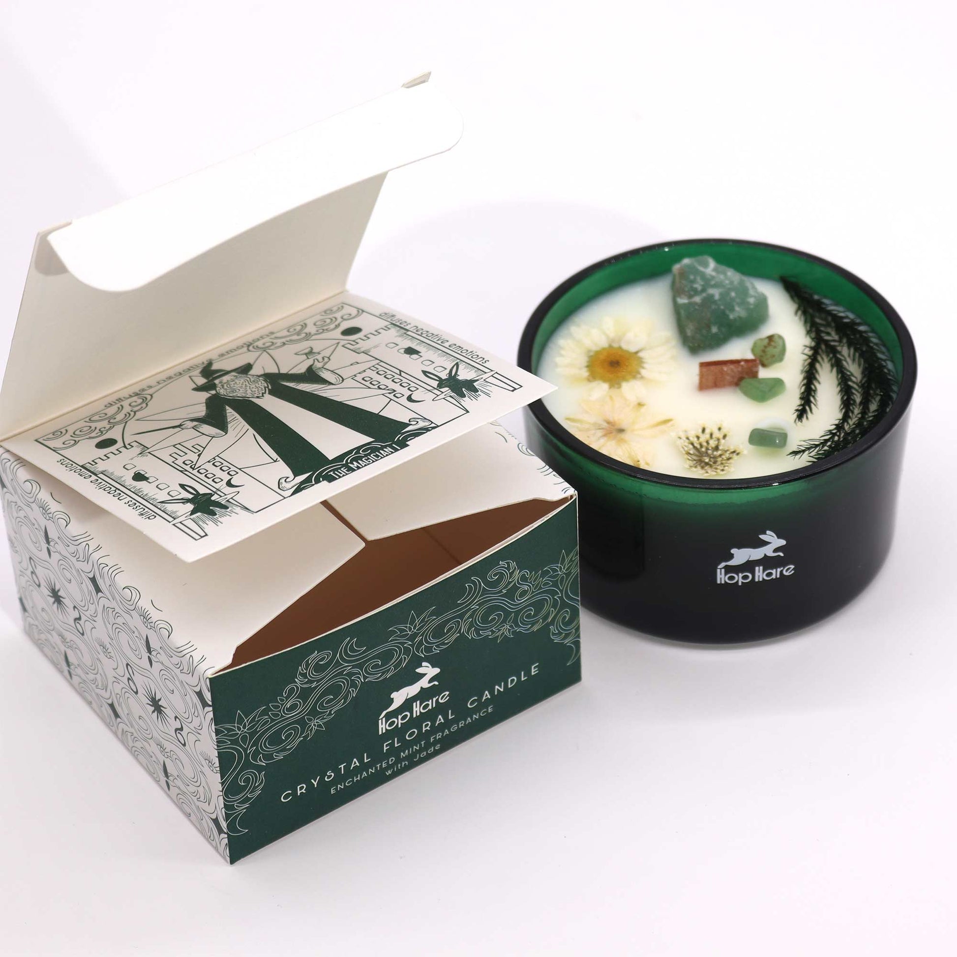 A candle sits next to its open box on a white surface. The candle has a dark green holder, white wax and decorative stones and dried flowers. The box says, "Hop Hare CRYSTAL FLORAL CANDLE ENCHANTED MINT FRAGRANCE with Jade," and has a drawing labeled, "THE MAGICIAN I."