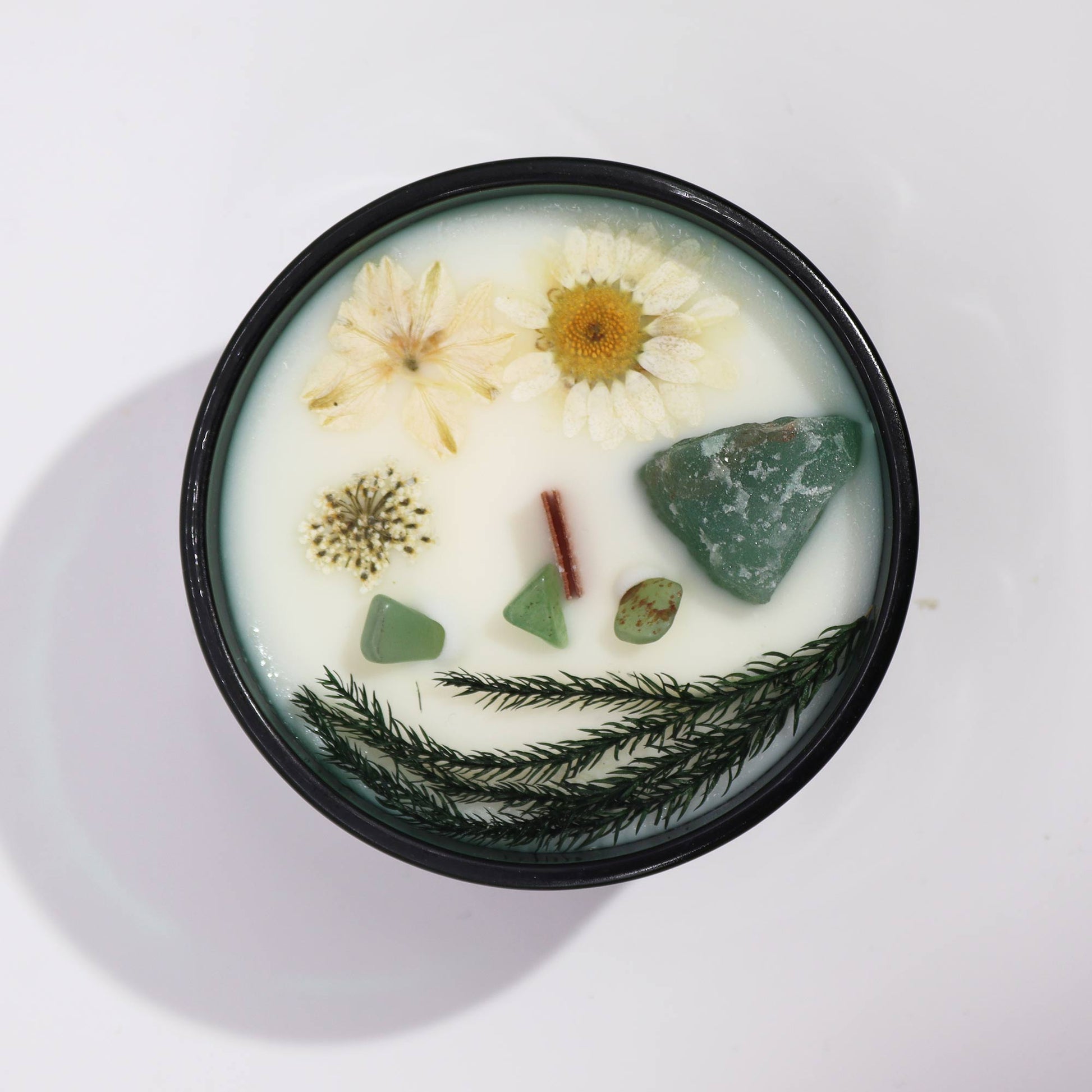 A black candle tin holds white wax embedded with dried flowers, green aventurine stones, a cinnamon stick, and evergreen sprigs on a white background.