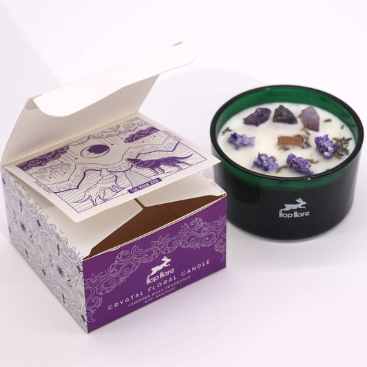 Green glass candle holds white wax topped with purple crystals and dried lavender, sitting beside an open purple-and-white decorative box on a white surface; Text: "Hop Hare", "CRYSTAL FLORAL CANDLE", "LAVENDER HILLS FRAGRANCE", "with Amethyst", "THE MOON XVI", "calm mind".