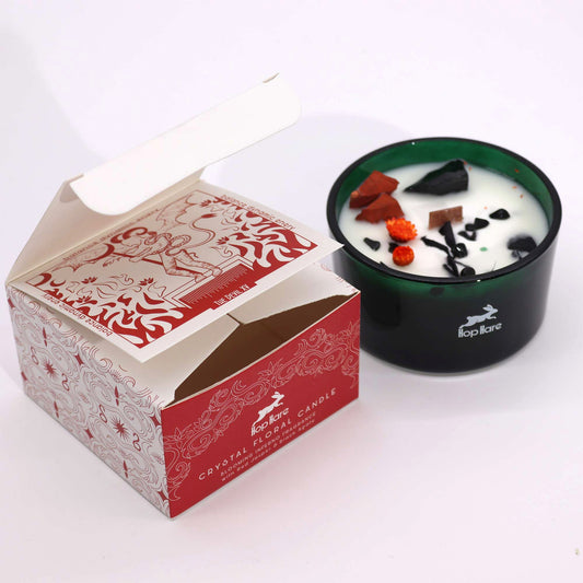 Green glass Hop Hare candle holds white wax topped with crystals and dried flowers while its red and white box lies open on a white surface Text: Hop Hare CRYSTAL FLORAL CANDLE THE DEVIL XV
