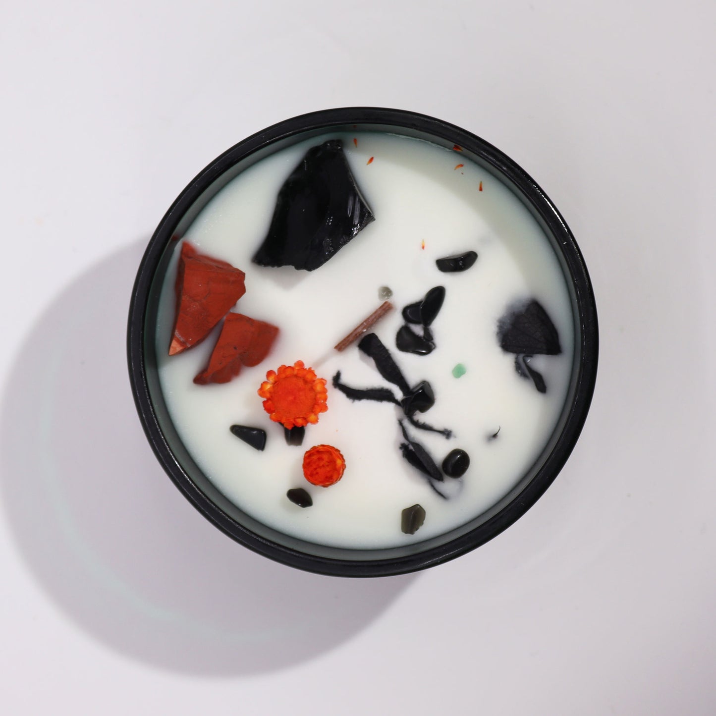 A black circular candle tin holds white wax, in which sit chunks of black and red rocks, dried orange flowers, and a cinnamon stick, on a white background.
