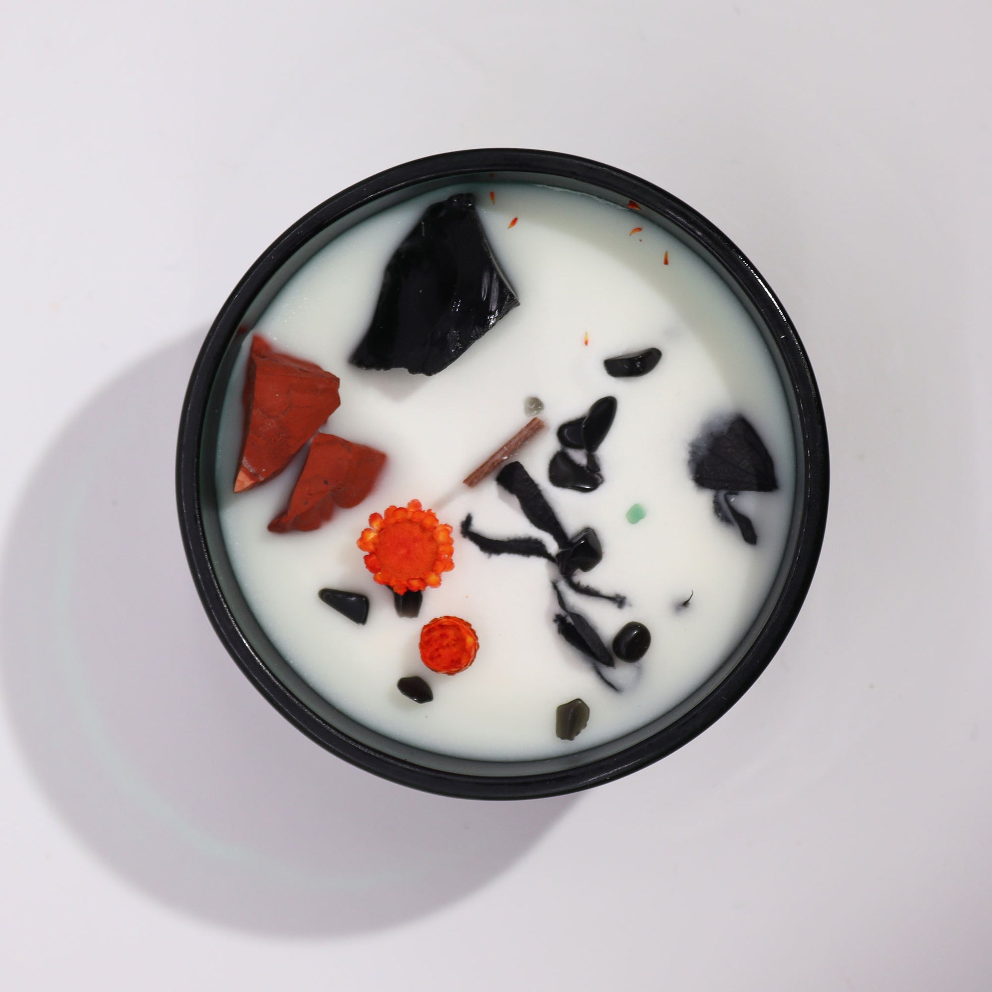 A black circular candle tin holds white wax, in which sit chunks of black and red rocks, dried orange flowers, and a cinnamon stick, on a white background.