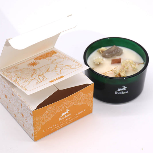 Green glass Hop Hare candle holds white wax topped with crystals and dried flowers beside its open orange-and-white box displaying an illustrated card; Text: Hop Hare CRYSTAL FLORAL CANDLE JASMINE GARDEN FRAGRANCE WITH ROSE QUARTZ STRENGTH releases energy and brings balance