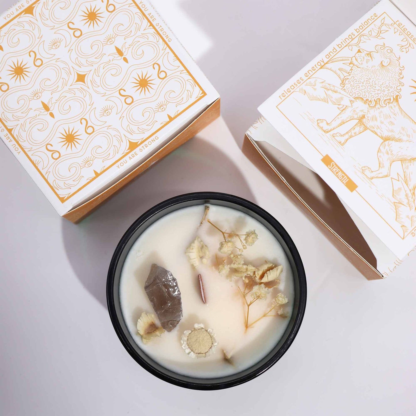 Candle in a black jar holds white wax topped with a smoky crystal and dried flowers (object) sitting between two white-and-gold patterned boxes on a pale surface (action/context); text: YOU ARE STRONG; release energy and brings balance; STRENGTH
