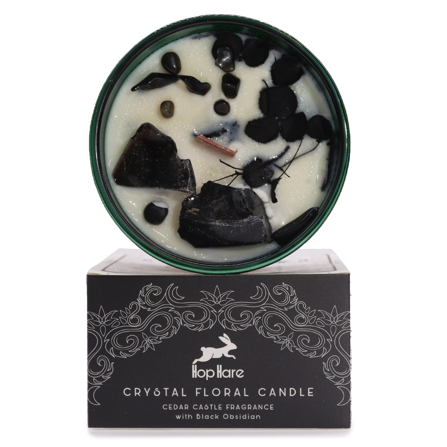 A green-rimmed candle contains wax and black obsidian stones and pieces. The candle sits atop its black packaging, which reads, "Hop Hare CRYSTAL FLORAL CANDLE CEDAR CASTLE FRAGRANCE with Black Obsidian."