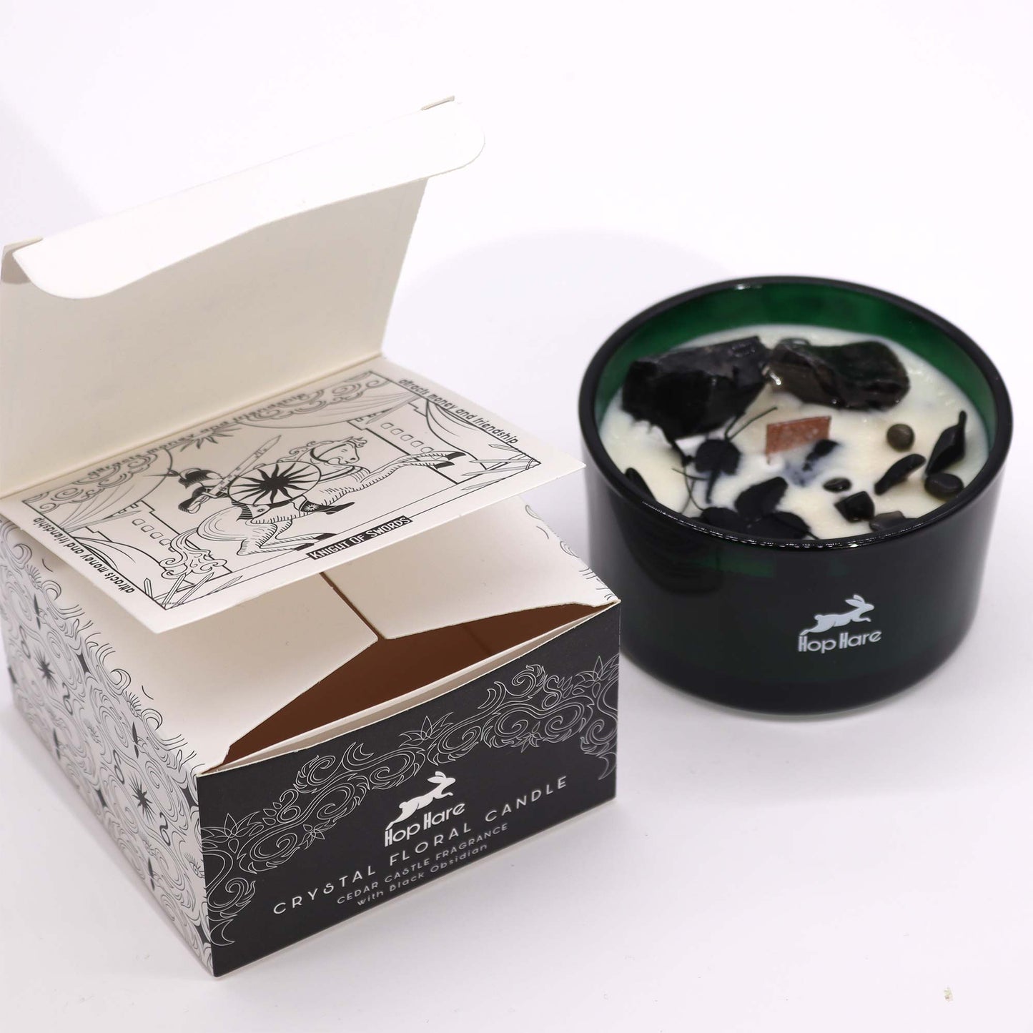 A partially opened box of "Hop Hare CRYSTAL FLORAL CANDLE CEDAR CASTLE FRAGRANCE with Black Obsidian" sits beside a green candle of the same brand, filled with wax and black obsidian on a white surface. Inside the box lid is text, "attracts money and friendship" and "KNIGHT OF SWORDS" with a drawing.
