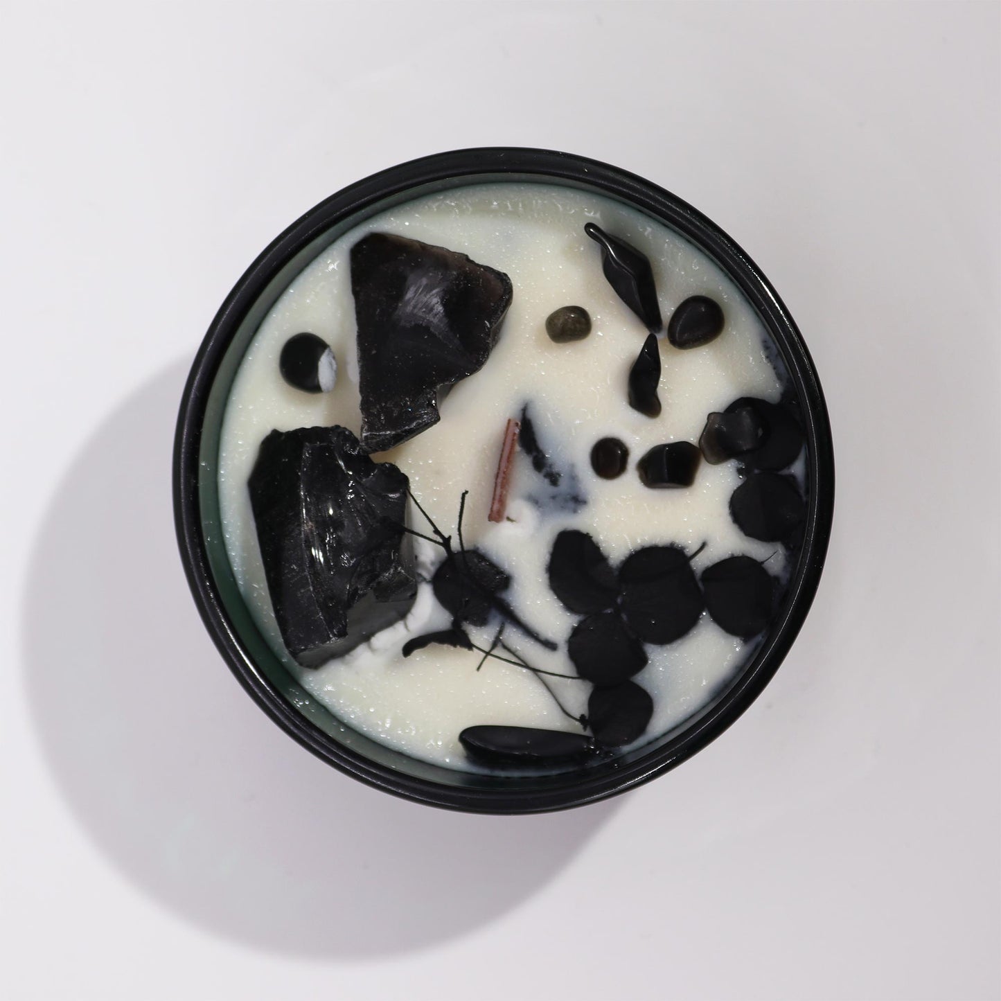 The candle contains black stones and botanicals in a creamy wax, held within a black circular tin, sitting on a plain white surface.