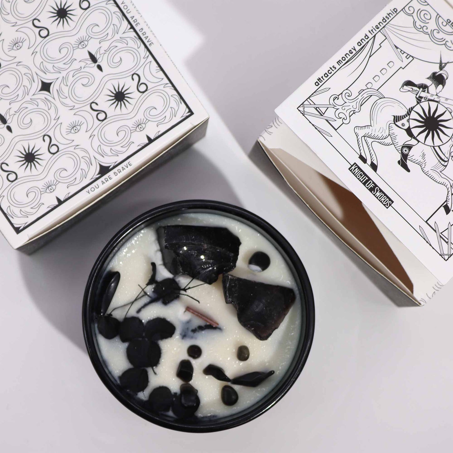 A black candle jar holds a white candle embedded with black stones, next to two boxes printed with black and white designs. The texts on the boxes include "YOU ARE BRAVE" and "attracts money and friendship KNIGHT OF SWORDS".