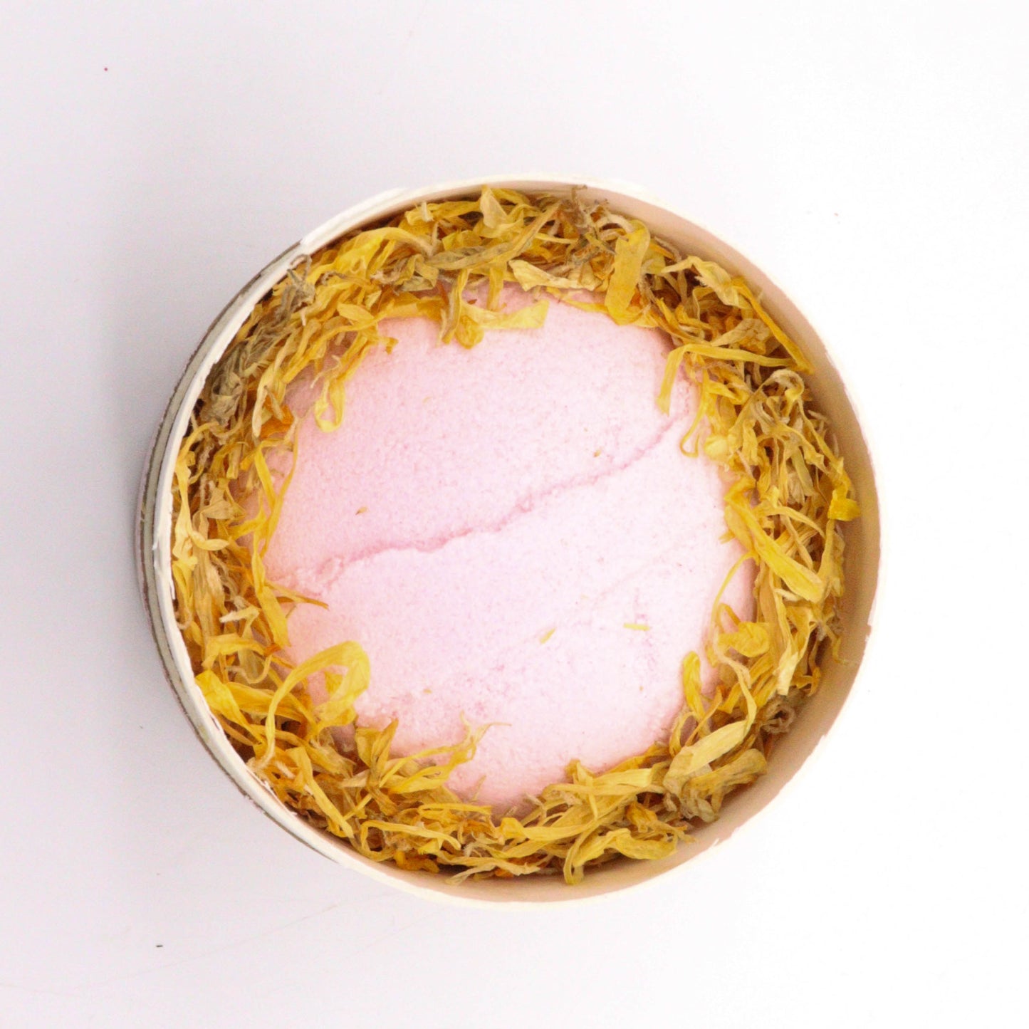 A round, pink bath bomb is nestled in a bed of dried yellow petals, placed in a cylindrical container on a white surface.
