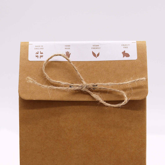 A brown paper package tied with twine rests against a plain background. A white label on the flap reads “MADE IN ENGLAND, HAND MADE, VEGAN FRIENDLY, CRUELTY FREE” with corresponding icons.