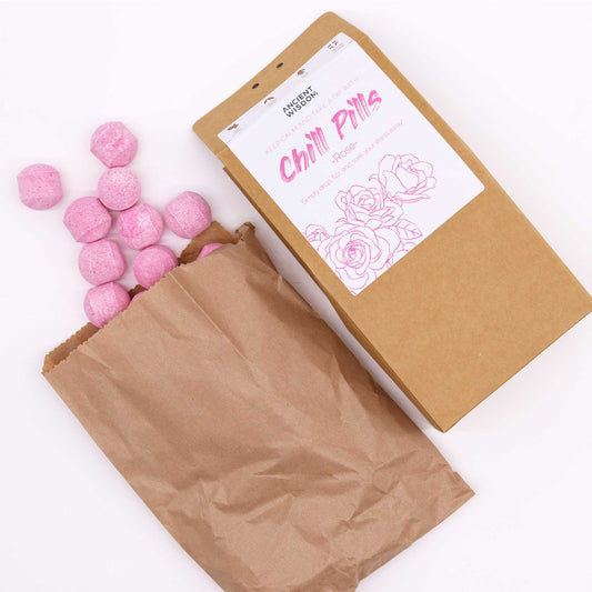 Pink bath bombs spill from a brown paper bag next to a cardboard box labeled "Ancient Wisdom Chill Pills - Rose." The label features rose illustrations and suggests calming use.
