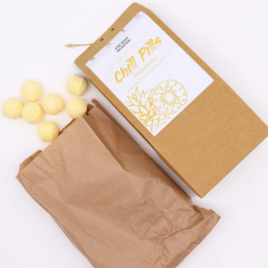 A brown paper bag dispenses yellow spherical tablets labeled "Chill Pills" with a pina colada theme from Ancient Wisdom, beside a labeled cardboard box on a white surface.