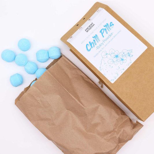 Blue bath bombs spilling from a brown paper bag beside labeled packaging that reads, "Ancient Wisdom, Keep Calm and Take a Dip with Chill Pills - Baby Powder, Simply drop, fizz and soak your stress away!"