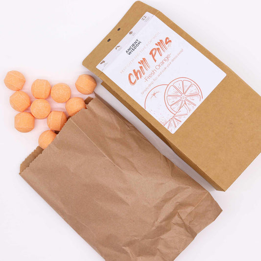 Orange bath bombs spill from a brown paper bag labeled "Ancient Wisdom Chill Pills Fresh Orange." The label features an orange slice illustration on a white background, set on a white surface.